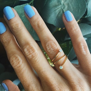May include: A gold-colored ring with a spiral design, featuring small, round, white gemstones. The ring is worn on a finger with light blue nail polish. The background includes green leaves.