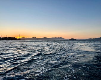 May include: A wide view of dark blue ocean water with visible ripples and white foam in the foreground, leading to a distant coastline under a gradient sky transitioning from deep orange at the horizon to a pale blue at the top of the frame.