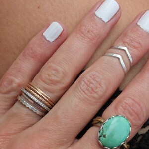 May include: A hand wearing a stack of silver, gold, and diamond rings on the ring finger, a silver chevron ring on the middle finger, and a turquoise ring on the pinky finger. The fingernails are painted white.