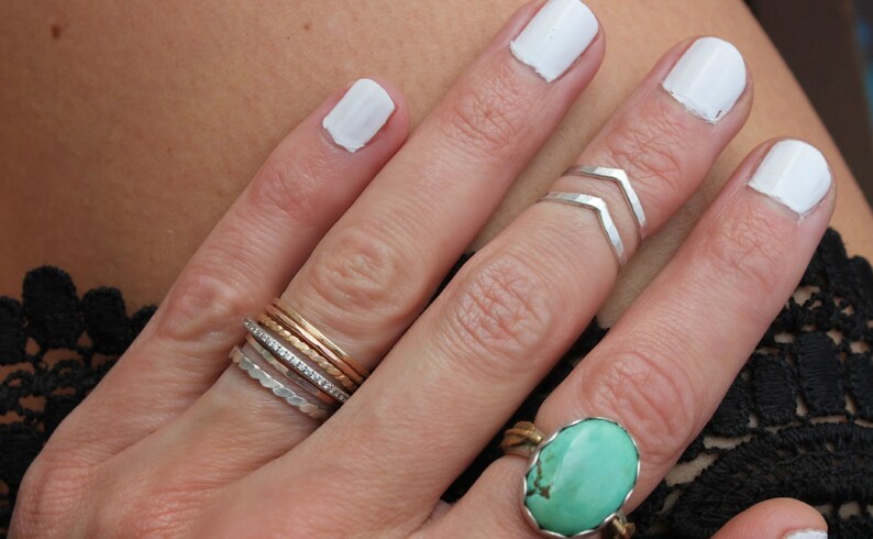 May include: A hand wearing a stack of silver, gold, and diamond rings on the ring finger, a silver chevron ring on the middle finger, and a turquoise ring on the pinky finger. The fingernails are painted white.