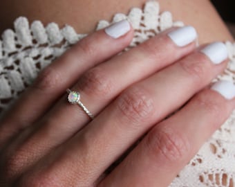 May include: A delicate silver ring featuring a small, round, iridescent opal stone set in a scalloped bezel. The ring band has a textured, twisted appearance, providing a subtle, detailed finish to the metallic surface of the jewelry piece.