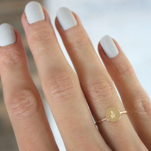 May include: A gold ring featuring a thin, textured band and a small, circular flat disc charm. The center of the disc is engraved with the lowercase letter w in a cursive script font. The ring is worn on a finger with light-colored nail polish.