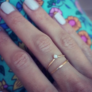 Pode incluir: Um anel de ouro com uma pedra de opala branca engastada no centro. O anel está em um dedo com esmalte de unha branco.