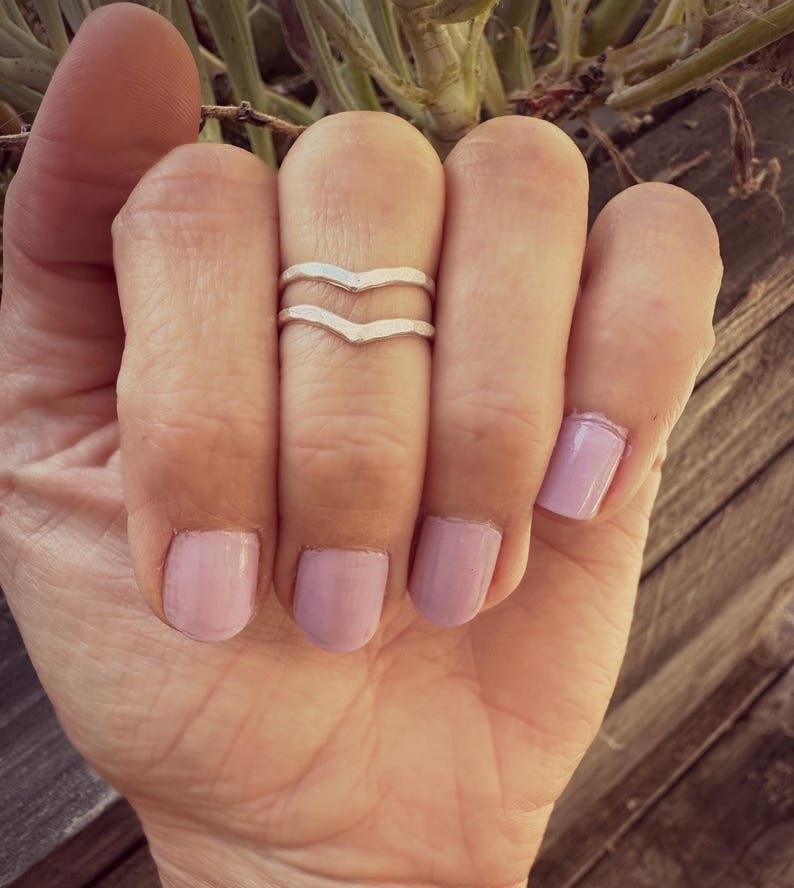 May include: A silver double chevron ring on a finger with light pink nail polish.