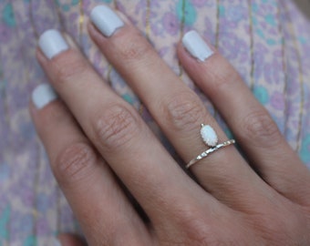 May include: A silver ring featuring a single, vertically oriented oval-shaped white opal stone set in a prong setting. The band of the ring has a textured, hammered finish, providing a distinct metallic surface detail across the thin metal band.