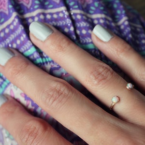 May include: A close-up of a hand wearing a delicate gold ring with two small white opal stones. The hand is resting on a purple and turquoise patterned fabric.