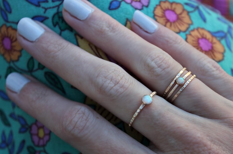 May include: Two gold rings with a white opal gemstone on a finger. The ring on the index finger is a simple band, while the ring on the middle finger is a triple band.