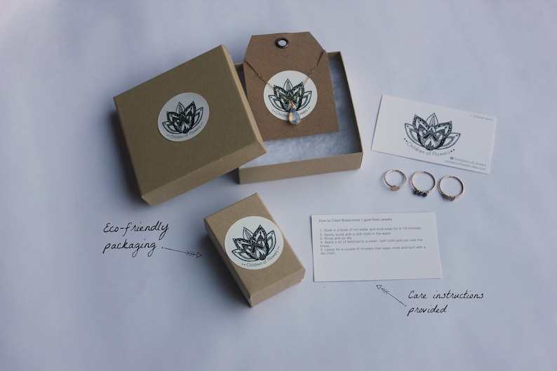 May include: A set of eco-friendly packaging for jewelry. The packaging includes a brown box with a white sticker that says "Garden of Flowers", a small brown box with a white sticker that says "Garden of Flowers", a white card with care instructions, and three gold rings with different stones.