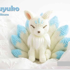 May include: A crocheted white fox with blue-tipped tails and ears, and closed eyes. The fox is sitting on a white surface. The text "Fuyuko The Kitsune" is in the upper left corner.