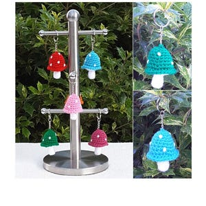 Op de afbeelding: Een display van handgemaakte gehaakte paddenstoel sleutelhangers in verschillende kleuren, waaronder rood, blauw, roze, groen en bordeaux. Elke paddenstoel heeft witte stippen en een witte steel. De sleutelhangers worden getoond op een zilveren metalen standaard.