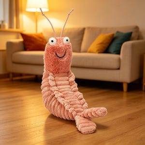 Puede incluir: Un peluche rosa con forma de camarón, con ojos grandes y redondos y una boca sonriente. El juguete tiene una textura similar a la pana y antenas largas y finas. Está sentado en un suelo de madera frente a un sofá.