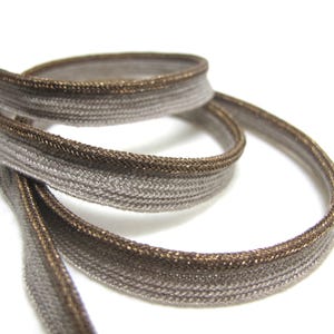 May include: Close-up of a coiled, metallic ribbon with a woven texture. The ribbon is primarily a light grey colour, with a darker, bronze-coloured trim along the edges. The ribbon is against a white background.