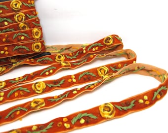 May include: Rust-colored velvet ribbon with embroidered yellow roses, green leaves, and small yellow dots. The ribbon is rolled up and also shown unrolled, displaying the floral pattern. This decorative ribbon is suitable for crafts and embellishments.