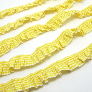 May include: Close-up of several strands of yellow and white gingham ruffle trim. The trim has a ruffled edge and is made of a woven fabric. The trim is ideal for sewing, crafting, and adding a decorative touch to various projects.