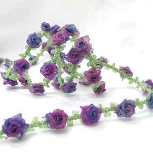 May include: A decorative floral ribbon featuring small, fabric roses in shades of purple and blue. The roses are attached to a green, vine-like base, creating a delicate and whimsical design. Perfect for crafts and embellishments.