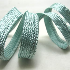 May include: A close-up of a length of light blue braided cord, coiled in loops. The cord has a tightly woven texture and a subtle sheen. The background is a plain white surface. This cord could be used for crafting or jewelry making.