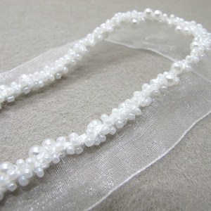 May include: A delicate white beaded trim with a sheer ribbon base. The trim features small, round, and oblong beads, creating a textured, elegant design. Ideal for embellishing bridal wear or craft projects.