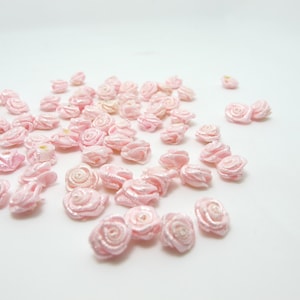 May include: A collection of small, light pink satin ribbon roses scattered on a white surface. The roses are tightly wound, with a spiral center and delicate petals. These miniature flowers are ideal for crafting and embellishments.