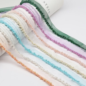 May include: Assortment of sheer ribbons in various colors, each adorned with a row of small, round beads. The ribbons include shades of green, purple, pink, blue, and peach. The word "STEIN" is visible on a white roll.