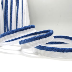 May include: White and blue striped elastic trim. Three coiled pieces of trim are in focus, with a roll of the same trim in the background. The trim has a white center with a blue, fuzzy border. The trim is likely used for sewing or crafting.