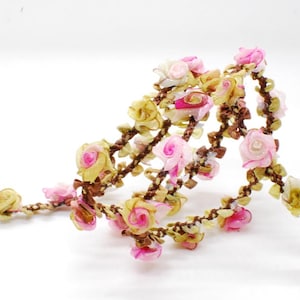 May include: A garland of small fabric roses in pink, yellow and white, strung on a brown, twisted cord. The roses have a delicate, handmade appearance, suitable for crafts or decorations.