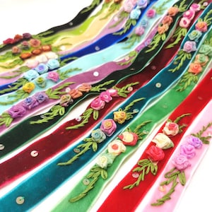 以下が含まれることがあります： 刺繍の花柄とスパンコールで飾られた、カラフルなベルベットリボンのコレクション。リボンはピンク、赤、緑、青など様々な色合いです。花の装飾には、小さなバラと緑の葉が使用されています。