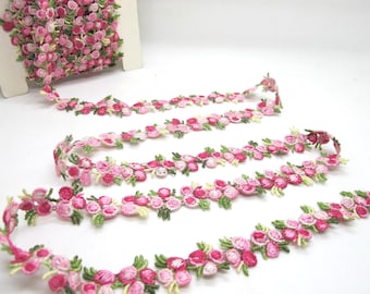 Op de afbeelding: Een rol roze bloemenrand met een delicaat ontwerp. De rand heeft kleine, geborduurde bloemen in roze en groene tinten, perfect voor het verfraaien van knutselwerkjes of kledingstukken. De rand staat op een witte achtergrond.