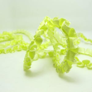 May include: Close-up of a length of light green decorative ribbon. The ribbon is ruffled and has a textured appearance. The ribbon is curled and twisted, creating a dynamic and visually appealing composition. The background is a soft, neutral white.