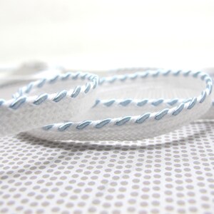 May include: Close-up of white ribbon with light blue stitching. The ribbon is coiled and lies on a patterned surface. The stitching adds a decorative element to the ribbon, which is likely used for crafts or embellishments.