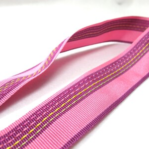Può includere: Un nastro rosa con un motivo a righe e cuciture decorative. Il nastro ha un colore di base rosa pieno con accenti viola e gialli. Il nastro è curvo e sembra essere fatto di un materiale simile al raso.