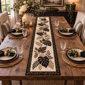 May include: A rectangular quilted table runner featuring a central cream-colored panel with a botanical pattern of dark brown and black leaves and vines, bordered by a black frame with gold-stitched vine details, centered on a wooden dining table.