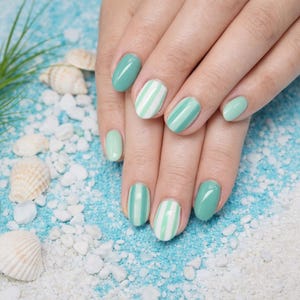 May include: Hands with manicured nails in shades of green and white. Some nails are solid green, while others feature vertical white stripes. The nails are oval-shaped and set against a beach-themed background with seashells and sand.