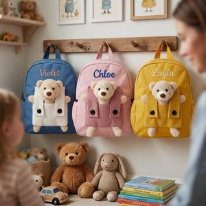 May include: Three children's backpacks in blue, pink, and yellow, each with a teddy bear in a pocket. The backpacks are personalized with the names Violet, Chloe, and Layla. Additional stuffed animals and books are visible.