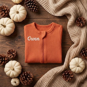 May include: An orange ribbed baby onesie with the name "Owen" embroidered in white. The onesie is zipped up and surrounded by white pumpkins, pine cones, and a beige knitted blanket on a wooden surface.