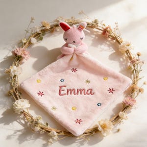 May include: A pink plush baby security blanket with a bunny head and the name "Emma" embroidered in red. The blanket is decorated with embroidered flowers and stars. The bunny has red ears and closed eyes.