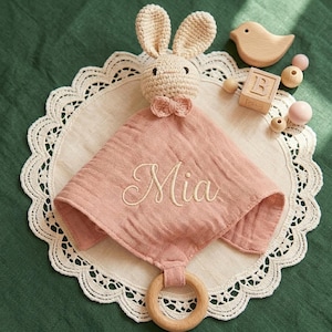 以下が含まれることがあります： かぎ針編みのウサギの頭が付いたベビー用コンフォートブランケット、ピンクのモスリン布、木製のリング。布には「Mia」という名前が刺繍されています。追加の木製おもちゃには、鳥、ビーズ、文字「B」のブロックが含まれています。