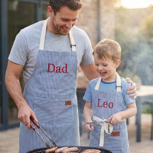 May include: Two aprons with blue and white stripes. One apron has the word "Dad" embroidered in red, and the other has "Leo". Both aprons have a small pocket and a tag that says "Apron".