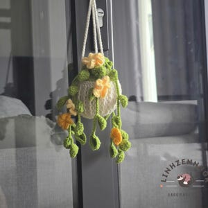Puede incluir: Un macetero colgante hecho a mano, tejido a ganchillo, con una maceta blanca adornada con flores amarillas y hojas verdes. El macetero está suspendido por un cordón blanco. El texto "LINHZENNNGR." y "HANDMADE." es visible.