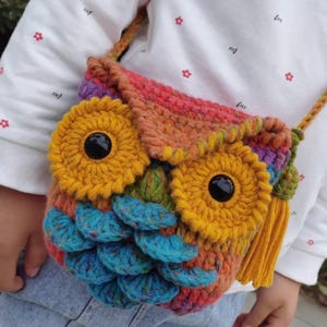 May include: A colorful, handmade crochet owl bag with yellow eyes, blue feathers, and a yellow tassel. The bag features a long strap and is made with various shades of yarn, including orange, pink, and purple.