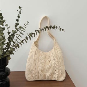 May include: Cream-colored knitted shoulder bag with a cable knit design on the front. The bag has a rounded shape and a single shoulder strap. The bag is made of a soft, textured yarn and is suitable for carrying everyday items.
