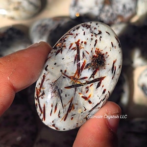 May include: An oval-shaped polished gemstone with a translucent white base featuring embedded needle-like inclusions in shades of dark brown, reddish-orange, and black. The text Ultimate Crystals LLC is printed in white on the lower right side.