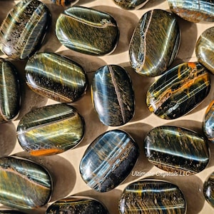 May include: A selection of polished tiger eye stones. The stones display a mix of colours, including gold, brown, and blue, with a glossy finish. The stones are oval-shaped and vary in size. The image is well-lit, highlighting the stones' natural patterns.