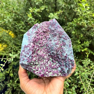 Op de afbeelding: Een ruw mineraal exemplaar met een cluster van diep magenta gekleurde kristallen ingebed in een getextureerde, lichtblauw-groene matrix. De steen heeft een onregelmatige, hoekige vorm met een ruwe, kristallijne oppervlaktestructuur.