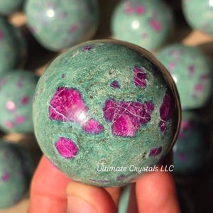 Peut inclure: Gros plan sur une sphère verte avec des inclusions roses et rouges. La sphère est tenue dans une main, avec d'autres sphères similaires en arrière-plan. Le texte "Ultimate Crystals LLC" est visible sur la sphère.