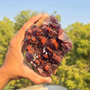 May include: A large, raw amethyst geode with deep red and purple crystalline structures. The geode is held in a hand, showcasing its natural, unpolished beauty. The colours range from dark red to a rich purple, with some areas appearing almost black.