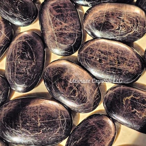 Puede incluir: Un conjunto de piedras negras pulidas, de forma ovalada, con intrincados patrones de vetas de color claro. Las piedras tienen un acabado brillante y están dispuestas juntas. El texto "Ultimate Crystals LLC" es visible.