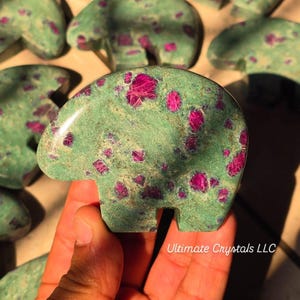 Könnte beinhalten: Eine grüne Edelstein-Bärenfigur mit rosa Einschlüssen. Der Stein ist in Form eines Bären geschnitzt. Der Text "Ultimate Crystals LLC" ist sichtbar. Der Bär wird in einer Hand gehalten.