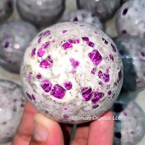 May include: A collection of gemstone spheres. The foreground sphere is white with vibrant magenta crystal inclusions. Other spheres are visible in the background, with a similar color palette. The spheres are held in a hand. The text "Ultimate Crystals LLC" is visible.