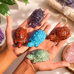 May include: A collection of gemstone turtle figurines in various colors, including purple, red, blue, orange, and green. The turtles are held in open hands, showcasing their detailed craftsmanship and unique textures. The background includes natural elements.