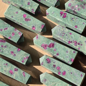 May include: A collection of green ruby zoisite obelisks. Each obelisk is rectangular with a pointed top and features vibrant pink ruby inclusions. The stones are arranged on a light brown surface, showcasing their natural beauty.